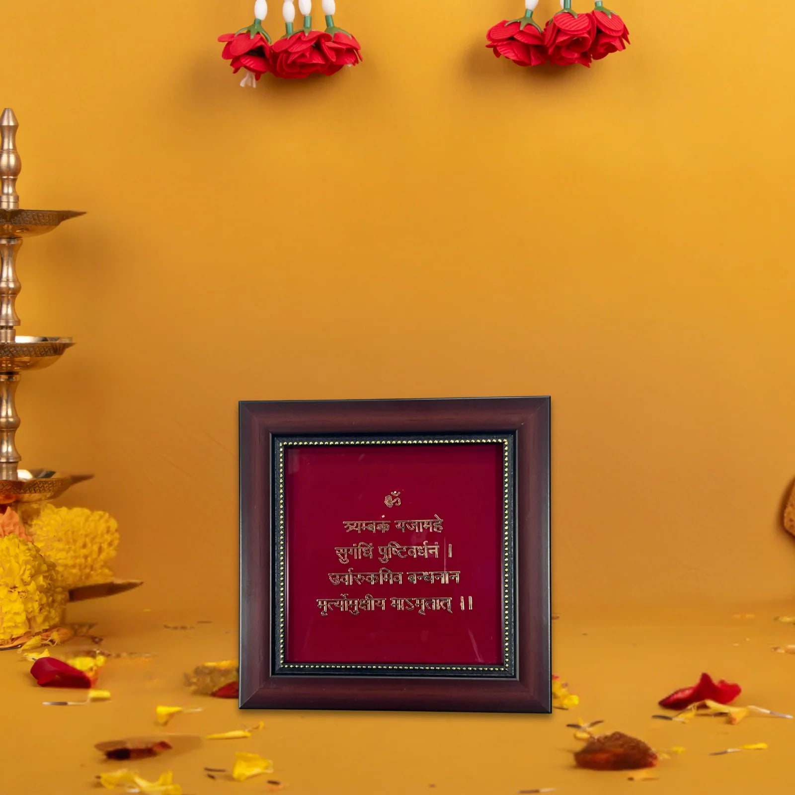 A wooden photo frame with a deep red background and golden text of - Mahamrityunjaya Mantra is placed on a surface. The text inside the frame is in Devanagari script. The frame is set against a yellow wall, with red flower garlands hanging above it. There are also some scattered flower petals and a brass lamp visible on the left side of the image.