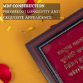 A brown MDF frame with a gold-beaded inner border, holding a red-backed panel with Hindi script in gold lettering, likely a Hindu mantra, viewed at an angle on a yellow surface with scattered yellow and red flower petals.