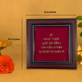 A brown wooden photo frame with a deep red backing contains the Mahamrityunjaya Mantra in gold-colored Sanskrit script. The mantra, which is dedicated to Lord Shiva, is visible in the center of the frame. The frame measures 6.5 inches by 6.5 inches, as indicated by text and dimension lines.