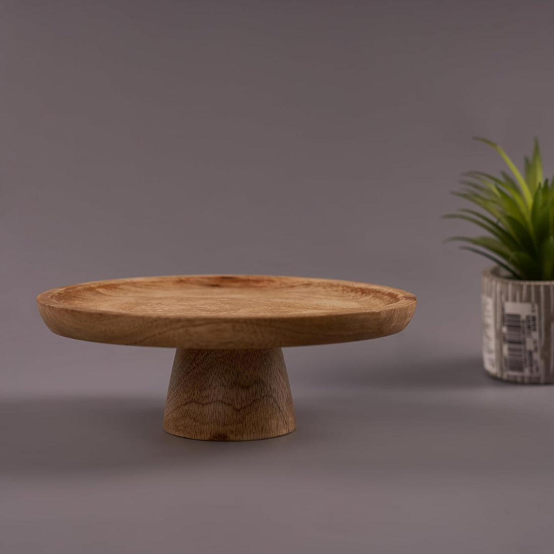 A mango wood cake stand with a rustic finish on a grey background, with a plant decoration to the side.