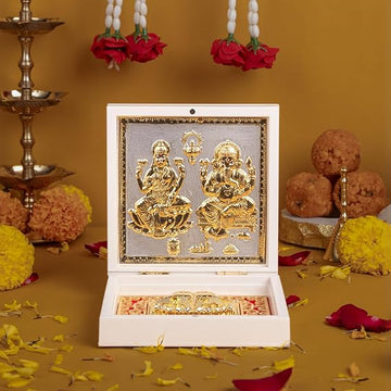 A white decorative box containing golden idols of Lakshmi and Ganesha, placed amidst marigold flowers, rose petals, traditional Indian sweets (ladoos), and brass diyas, likely set up for a Hindu festival or puja celebration.
