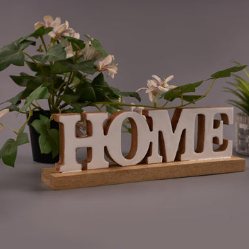 A wooden sign with the word 'HOME' displayed in capital letters, placed on a surface with a potted plant in the background.