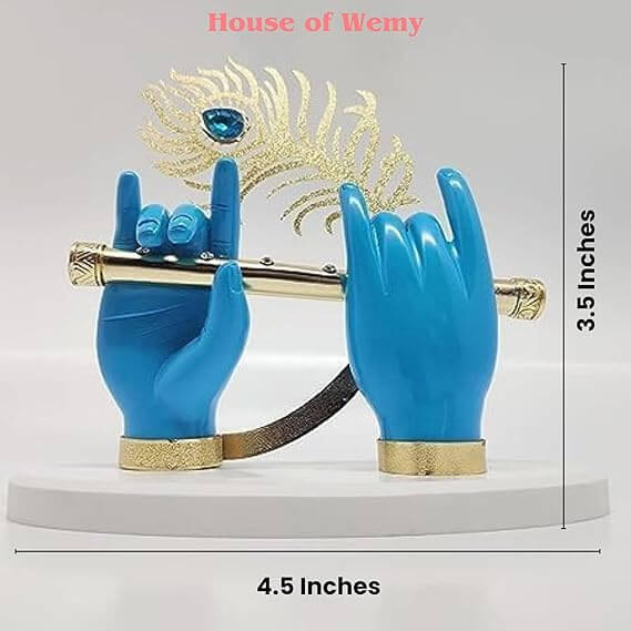 A decorative idol featuring two blue hands, likely representing Lord Krishna's, holding a golden flute. A golden peacock feather with a blue gem is positioned behind the hands, all resting on a white oval base. The image also displays text indicating the dimensions "3.5 Inches" and "4.5 Inches", along with the brand name "House of Wemy".