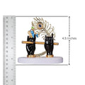 A close-up of a Lord Krishna idol depicting two black hands holding a golden flute, adorned with a golden and blue peacock feather, placed on a white base. A ruler on the left side shows the idol's height to be approximately 4.5 inches.