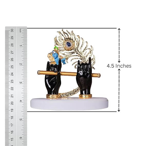 A close-up of a Lord Krishna idol depicting two black hands holding a golden flute, adorned with a golden and blue peacock feather, placed on a white base. A ruler on the left side shows the idol's height to be approximately 4.5 inches.