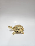A small, gold-plated turtle figurine, a Feng Shui decorative item, is shown on a simple white surface. The turtle has a detailed shell and a shiny finish.