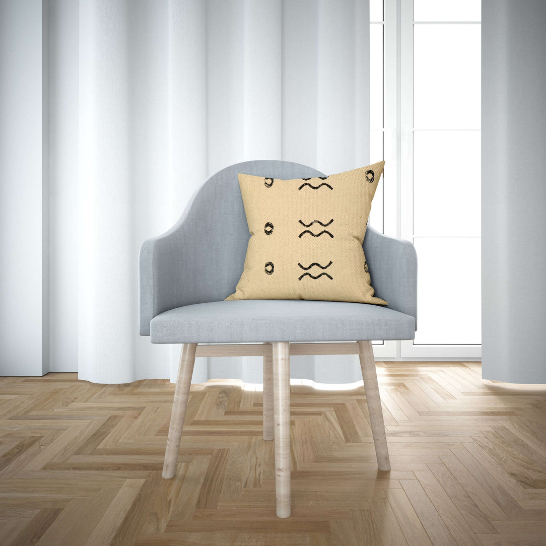 A cushion cover with a block print design displayed on a chair in a room with wooden flooring and white curtains.