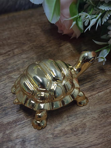 gold-plated turtle figurine, a Feng Shui decorative item, is shown on a rustic wooden surface. The turtle has a ribbed shell and is positioned in front of a soft-focus background of white and pink flowers.