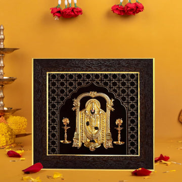 A gold-plated Lord Balaji photo frame with a dark, ornate border, featuring a detailed golden depiction of the deity, placed against a warm yellow-orange background with red flowers and petals, alongside traditional brass lamps.
