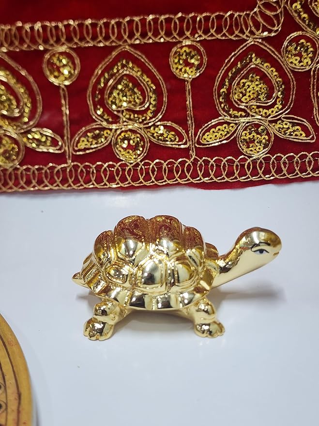 A close-up of a small, gold-plated turtle figurine, a Feng Shui decorative item, positioned on a white surface. The turtle has a detailed shell with segments and its head is turned slightly to the right. In the background, there is a red and gold embroidered fabric.