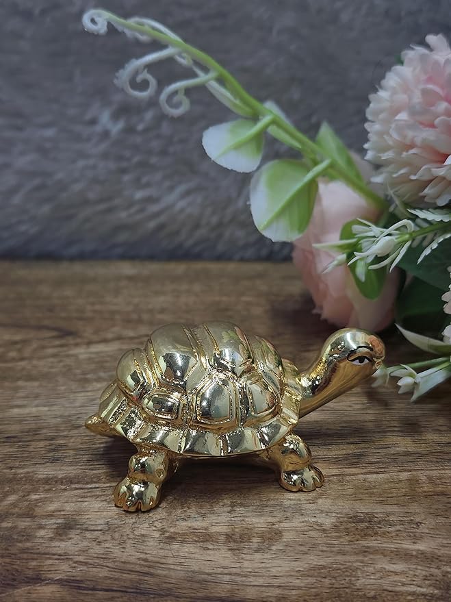A small, gold-plated turtle figurine, a Feng Shui decorative item, is shown on a rustic wooden surface. The turtle has a shiny, smooth finish and a detailed shell. In the soft-focus background, there is a floral arrangement with white and pink flowers.