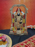 Gold-plated idol of Lord Tirupati Balaji (Venkateswara) standing within an ornate arch, set on a dark surface with a plate of traditional offerings and a red cloth in the foreground.