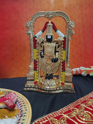 Gold-plated idol of Lord Tirupati Balaji (Venkateswara) standing within an ornate arch, set on a dark surface with a plate of traditional offerings and a red cloth in the foreground.