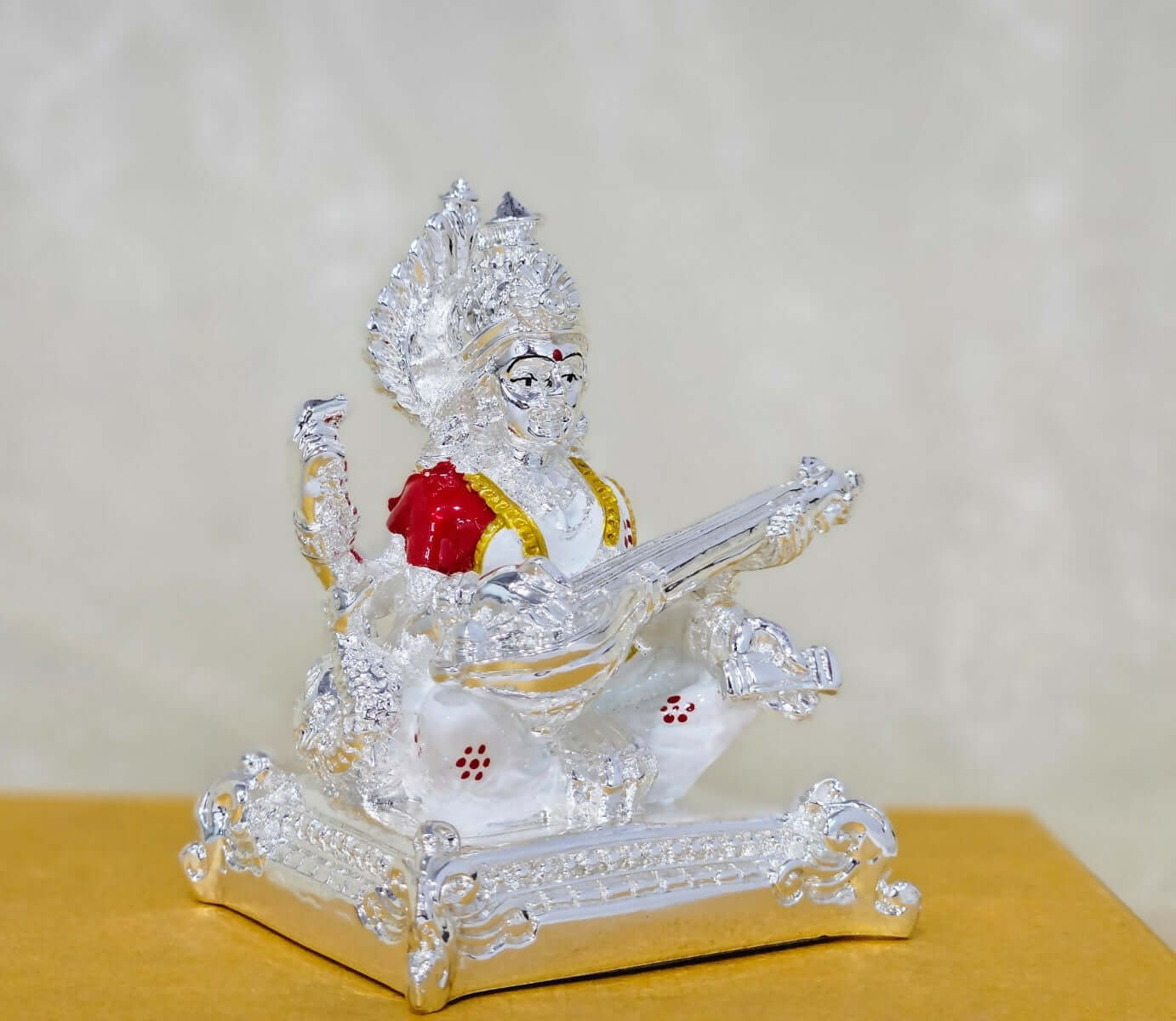 An image of a silver-plated idol of the Hindu goddess Saraswati seated and playing the veena, with intricate details and accents of red and gold.