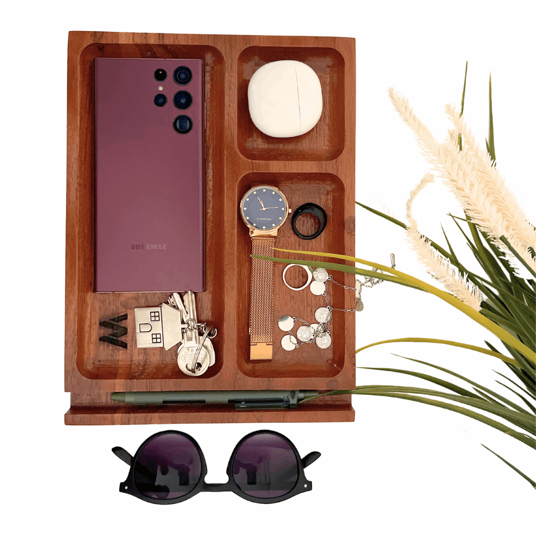 A wooden desk organizer tray with multiple compartments. The tray holds a purple smartphone, a watch, a ring, a necklace, and a small, smooth white stone. A pair of keys with a house-shaped keychain and a pen are also stored in the tray, which sits on a wooden base. A pair of sunglasses rests in front of the tray, and a decorative plant is on the right side.