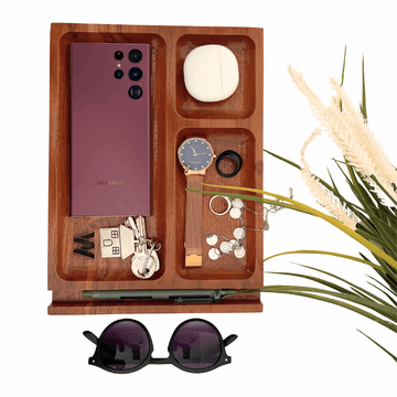 A wooden desk organizer tray with multiple compartments. The tray holds a purple smartphone, a watch, a ring, a necklace, and a small, smooth white stone. A pair of keys with a house-shaped keychain and a pen are also stored in the tray, which sits on a wooden base. A pair of sunglasses rests in front of the tray, and a decorative plant is on the right side.