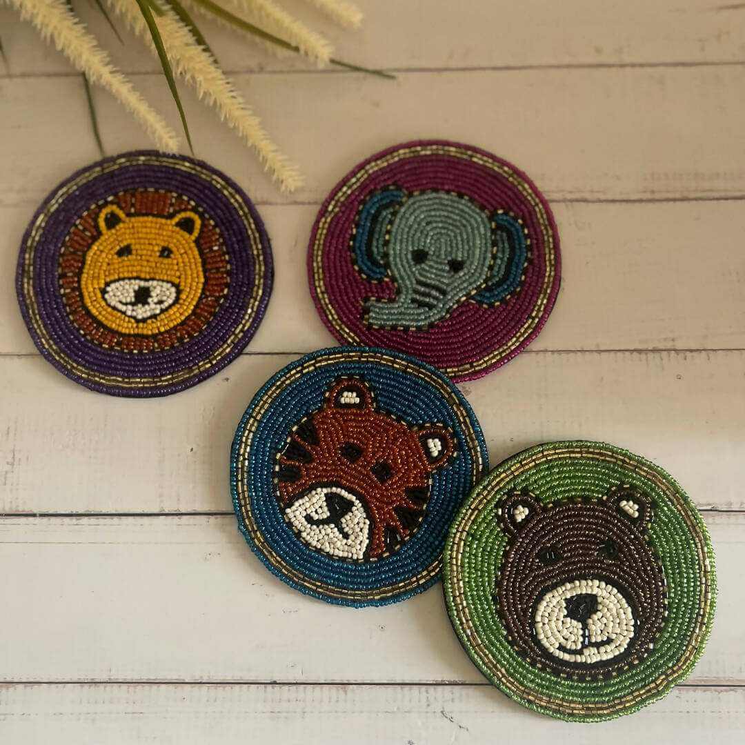 A collection of four Beaded Coasters - Safari Theme on a rustic wooden surface. Each coaster features the face of a different beaded animal: a yellow lion, a light blue elephant, a brown tiger, and a dark brown bear.