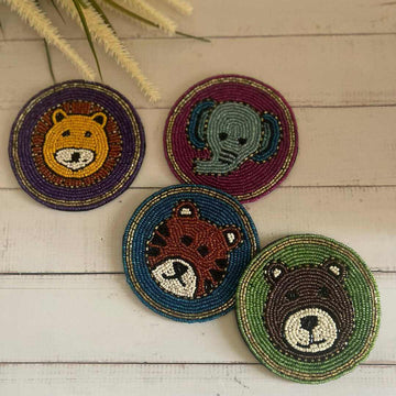 A collection of four Beaded Coasters - Safari Theme on a rustic wooden surface. Each coaster features the face of a different beaded animal: a yellow lion, a light blue elephant, a brown tiger, and a dark brown bear.