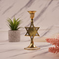 A gold-colored star-shaped brass candle holder displayed on a marble surface with a potted plant and decorative flowers in the background.