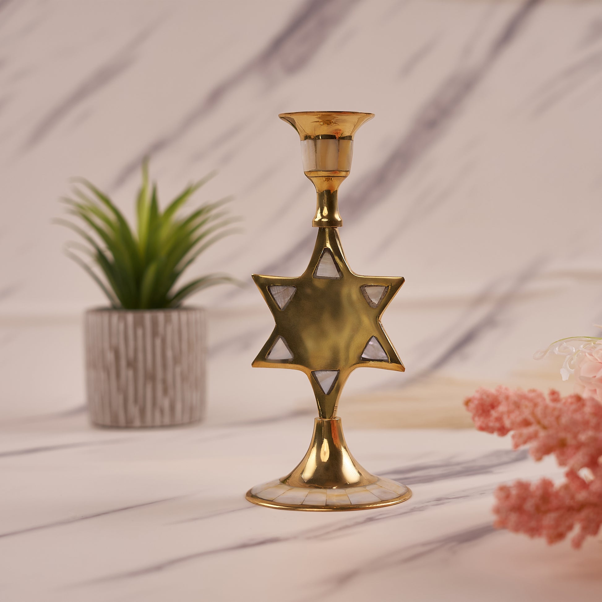 A gold-colored star-shaped brass candle holder displayed on a marble surface with a potted plant and decorative flowers in the background.