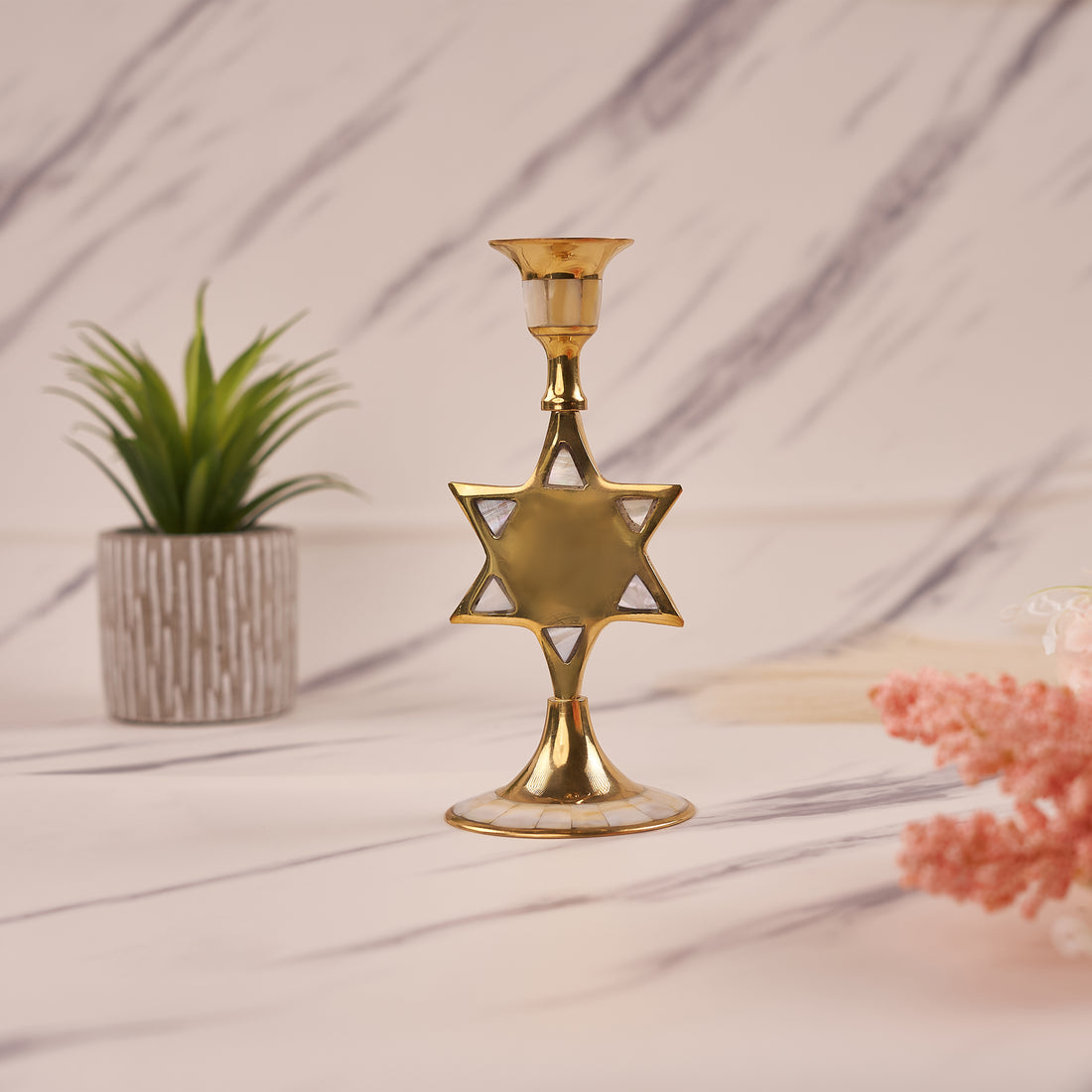 A gold-colored star-shaped brass candle holder displayed on a marble surface with a potted plant and decorative flowers in the background.
