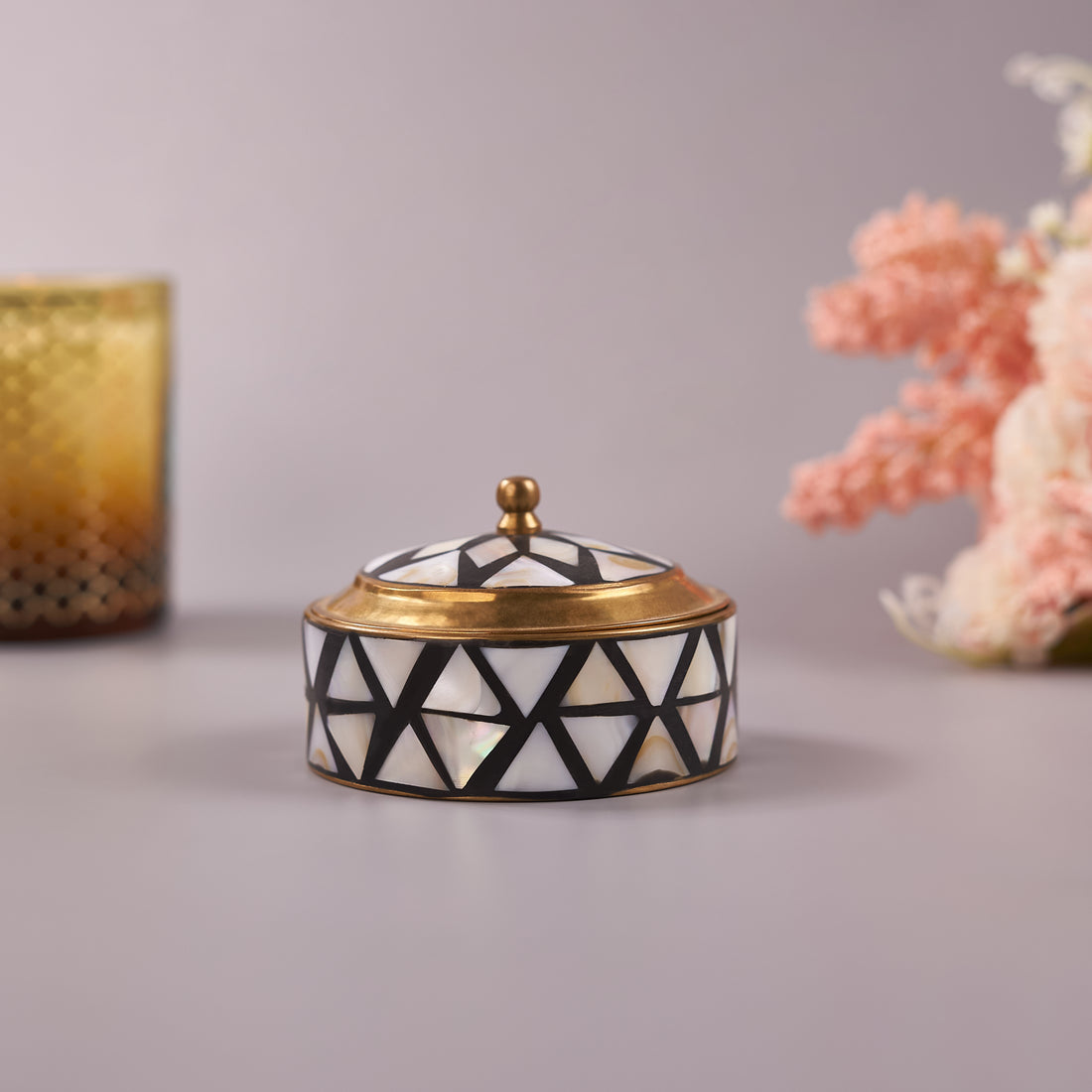 A small brass trinket box with mother-of-pearl inlay on a lid, placed on a flat surface with a floral arrangement in the background.