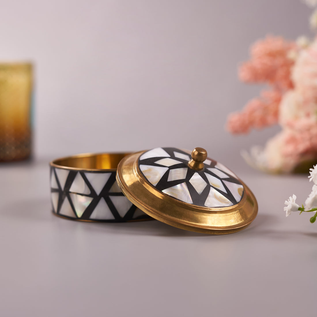 A small brass trinket box with mother-of-pearl inlay on a lid, placed on a flat surface with a floral arrangement in the background.