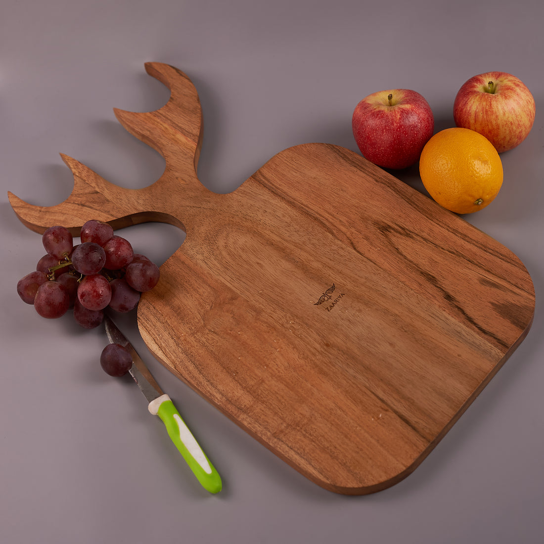 A wooden chopping board with a reindeer shape at the top, displayed with a knife and sliced fruits on it, against a grey background.