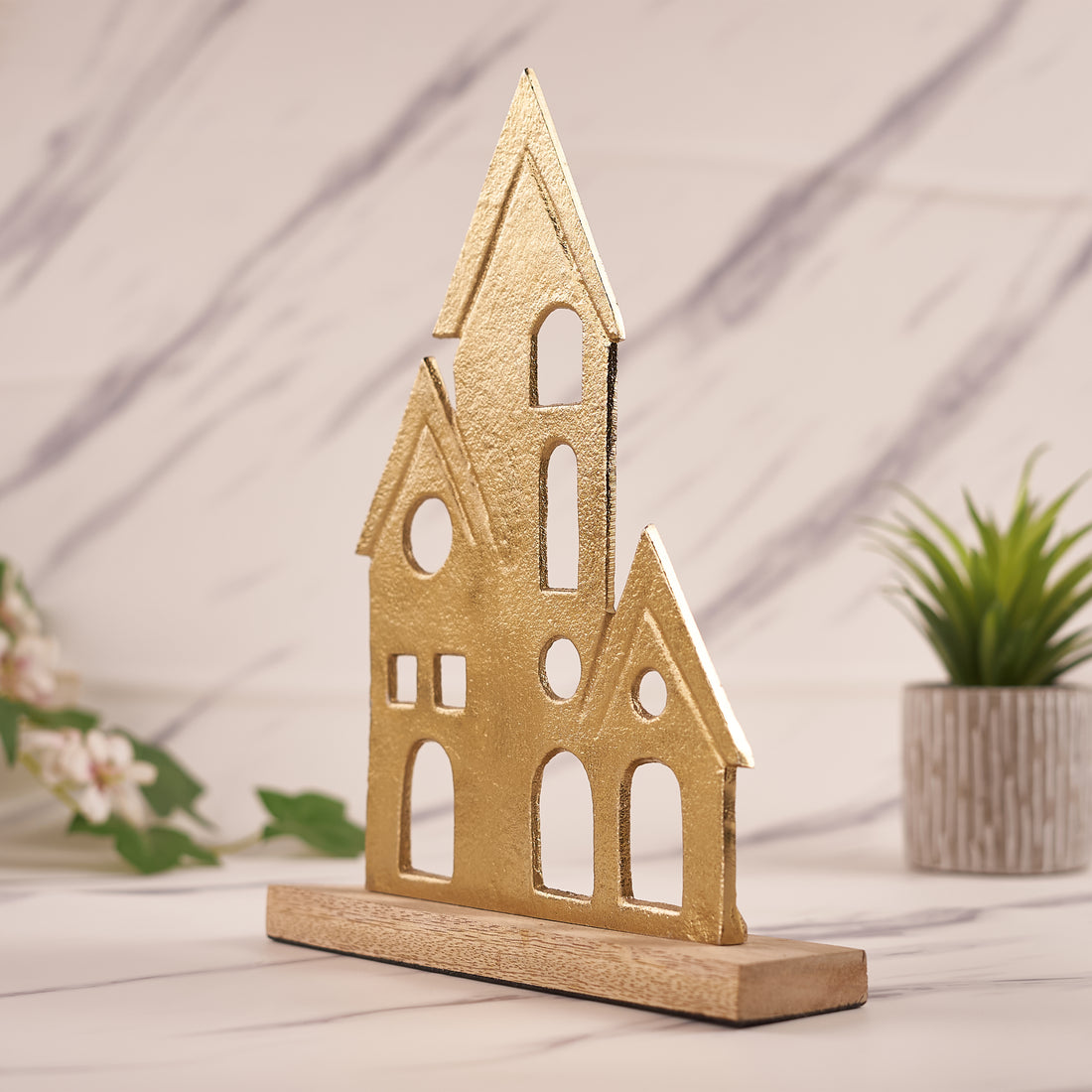 A brass house figurine with a wooden base, placed on a marble surface, with a plant decoration to the side.