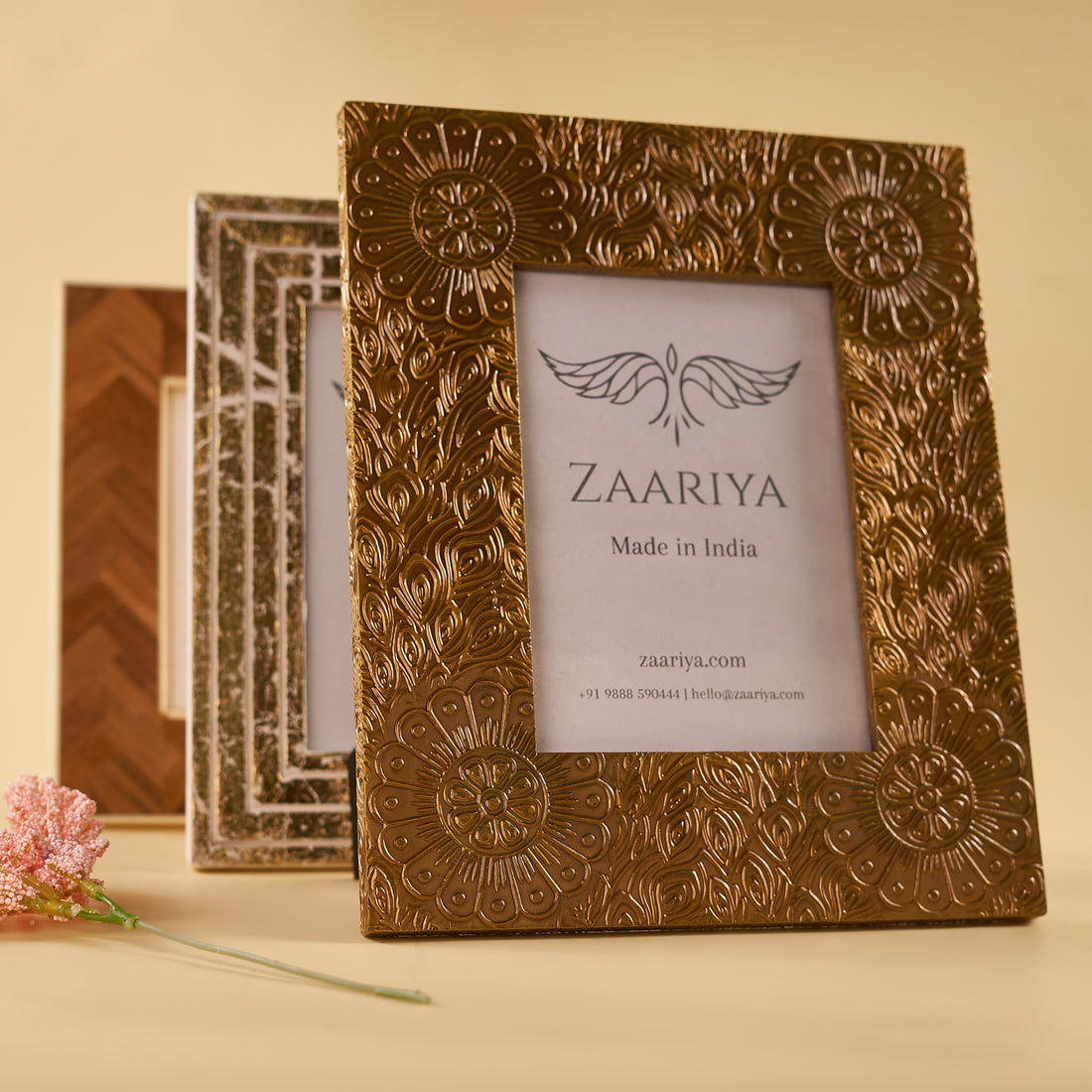 A wooden photo frame with a gold finish and floral patterns around the border, with a white mat and a picture of a house inside, placed on a yellow surface with a pink flower beside it.