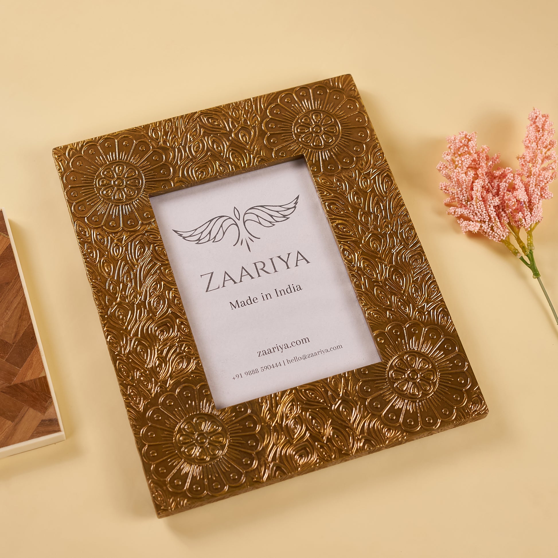 A wooden photo frame with a gold finish and floral patterns around the border, with a white mat and a picture of a house inside, placed on a yellow surface with a pink flower beside it.