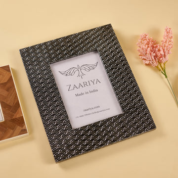 A wooden silver photo frame with a patterned design around the border, placed on a flat surface with a decorative item to the side.
