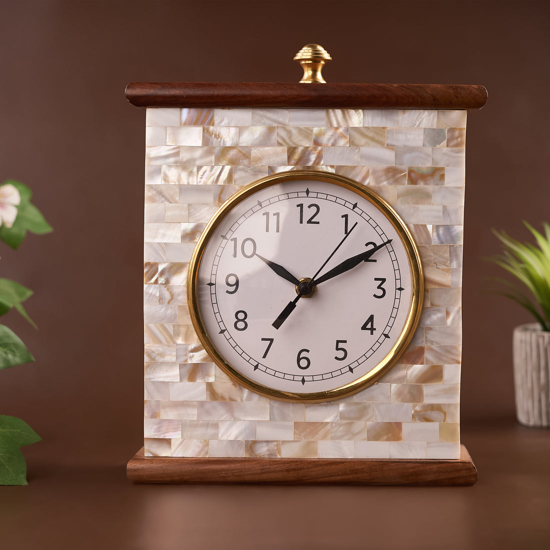 A Mother of Pearl table clock with a wooden frame, featuring a white clock face with black numerals, set against a brown background.
