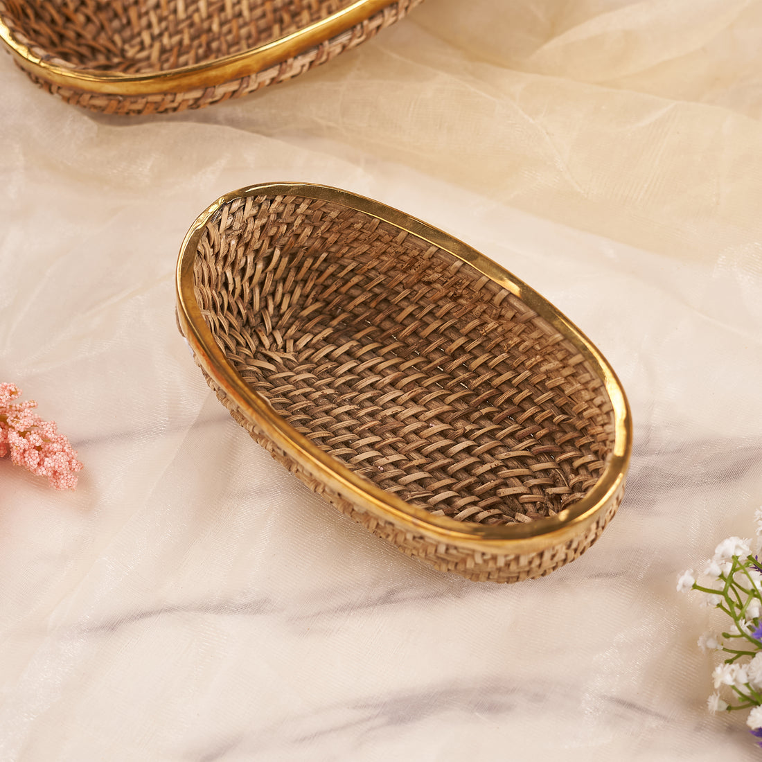 A long oval rattan basket with a beige color and a gold-colored rim, displayed on a soft, light background accompanied by small flowers.