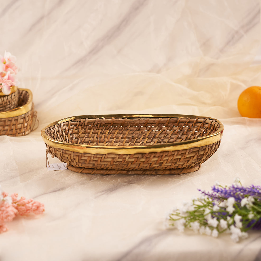 A close-up shot of a Large Oval Rattan Basket on a marble-patterned surface. The basket features a tight, woven pattern of natural rattan and is framed by a sleek, golden metallic rim. In the background, another similar basket is partially visible, while some delicate white and pink flowers are in the foreground.
