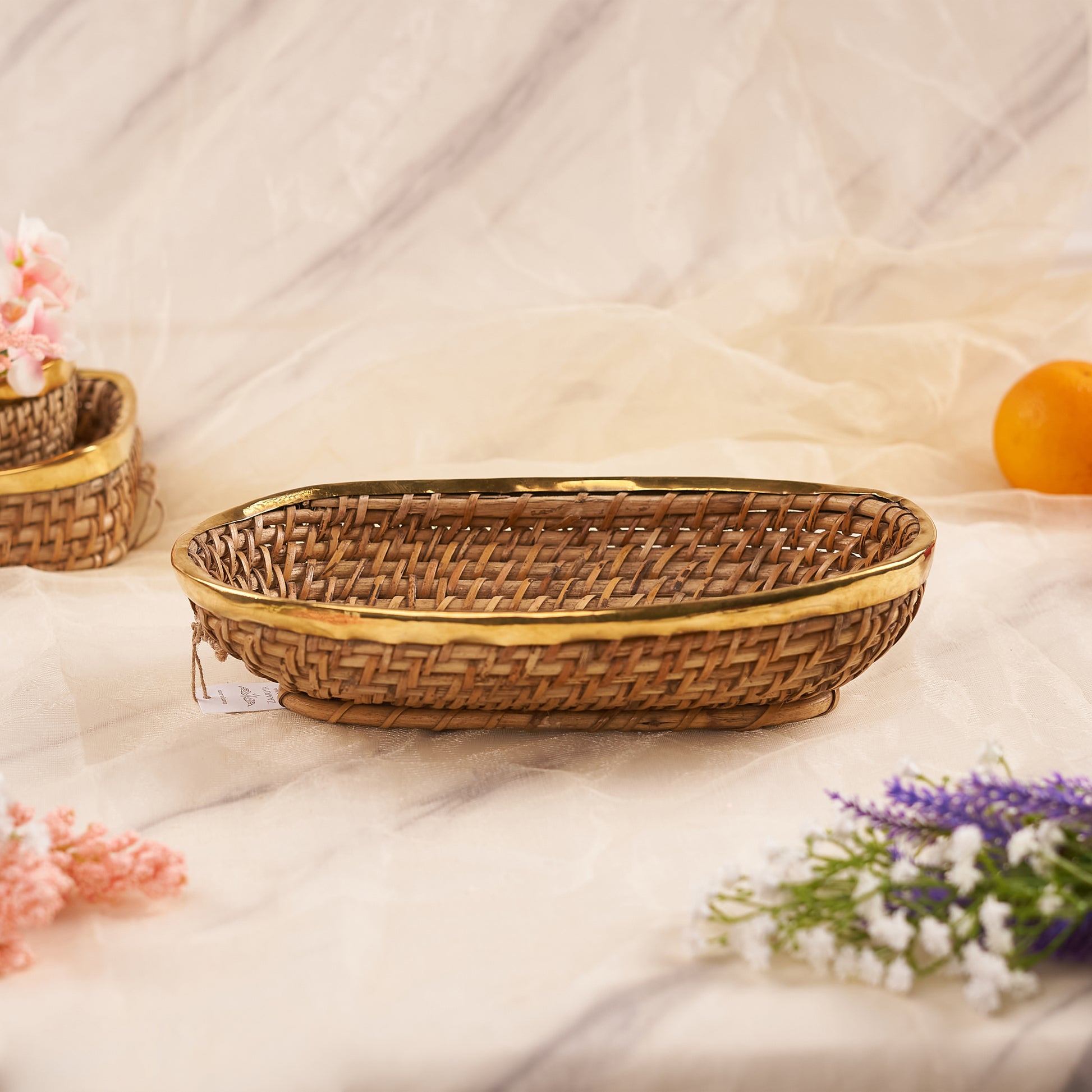 A Large Oval Rattan Basket is shown on a light-colored, marble-patterned surface. The basket has a tight, woven pattern of natural rattan and is finished with a prominent, sleek golden metallic rim. In the background, there is a similar smaller basket, an orange fruit, and some flowers, while delicate pink and purple flowers are visible in the foreground.