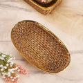 A close-up shot of a Large Oval Rattan Basket on a marble-patterned surface. The basket features a tight, woven pattern of natural rattan and is framed by a sleek, golden metallic rim. In the background, another similar basket is partially visible, while some delicate white and pink flowers are in the foreground.