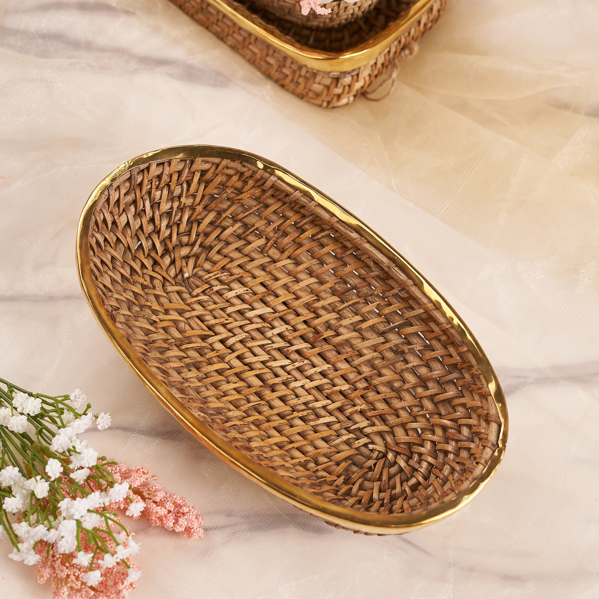 A close-up shot of a Large Oval Rattan Basket on a marble-patterned surface. The basket features a tight, woven pattern of natural rattan and is framed by a sleek, golden metallic rim. In the background, another similar basket is partially visible, while some delicate white and pink flowers are in the foreground.