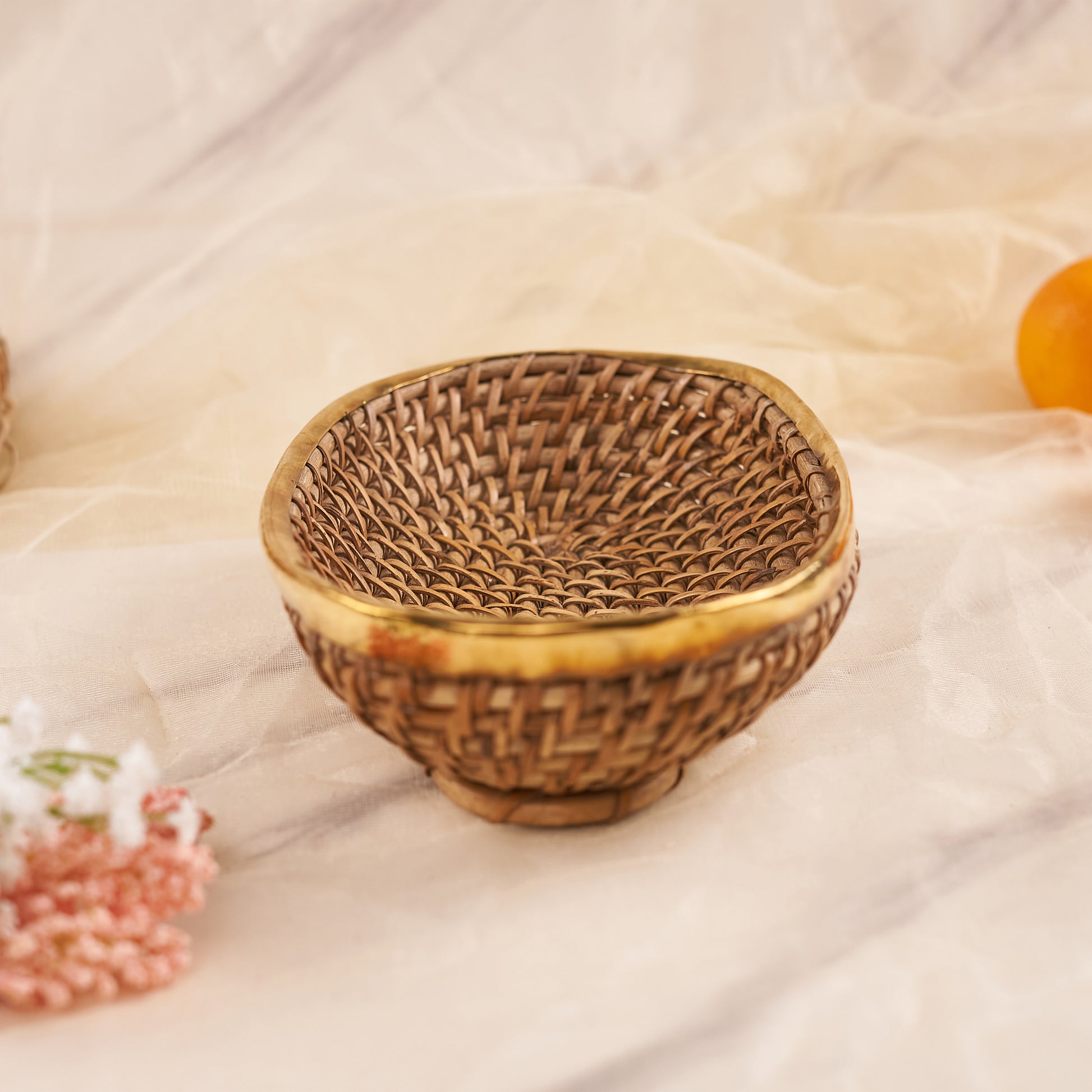 A small, round rattan basket with a tight weave and a golden metallic rim is shown on a light-colored, marble-patterned surface. While this particular basket is small and round, it is a variation of a Large Oval Rattan Basket and has a similar woven pattern and golden rim. A small pink and white floral arrangement is partially visible in the foreground, and an orange fruit is visible in the background.