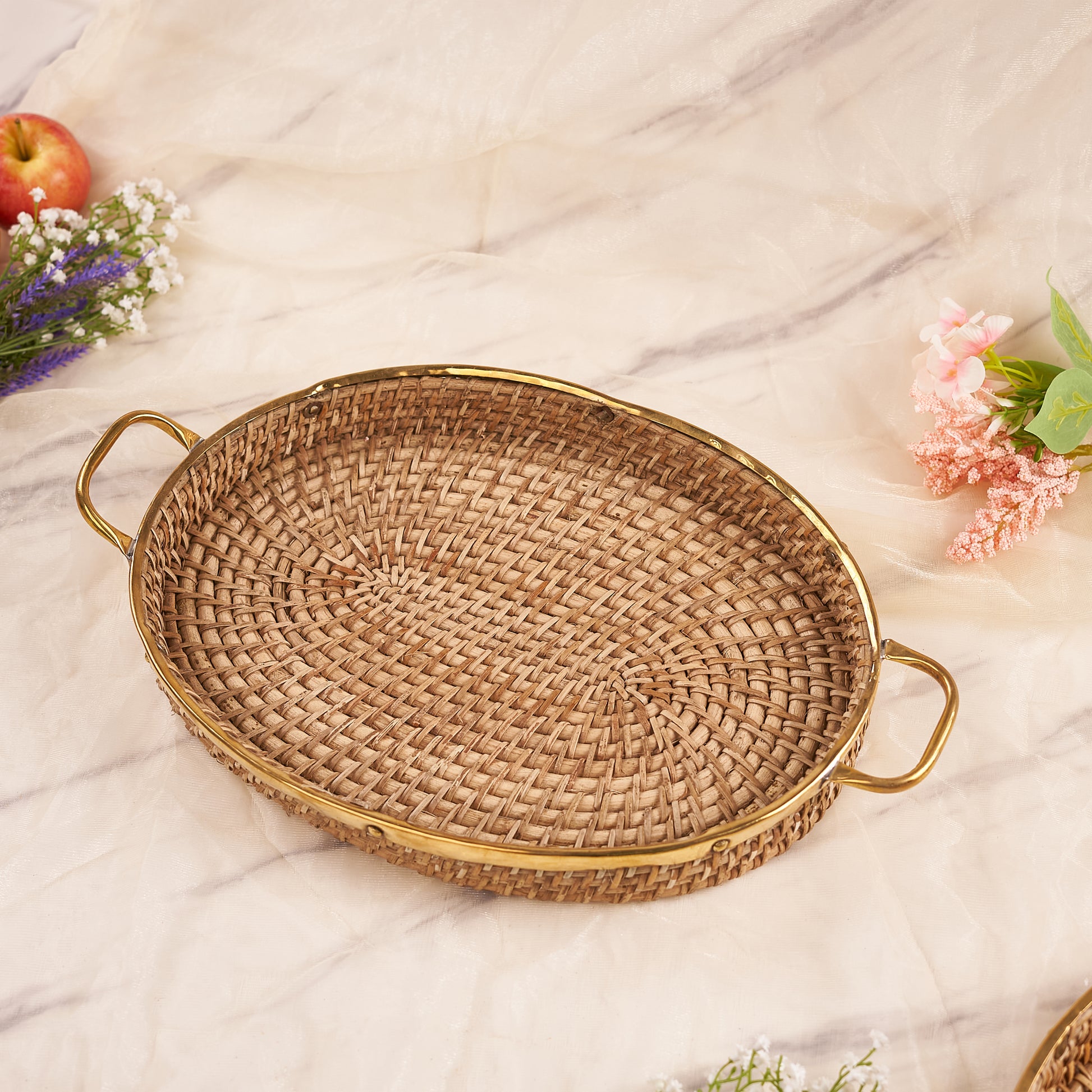 An oval rattan tray with handles, displayed on a marble surface surrounded by flowers and fruits.