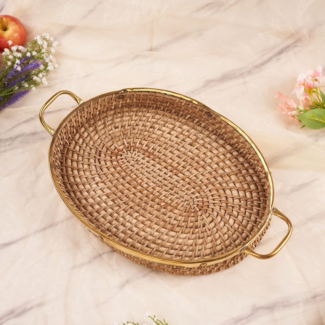 An oval rattan tray with handles, displayed on a marble surface surrounded by flowers and fruits.