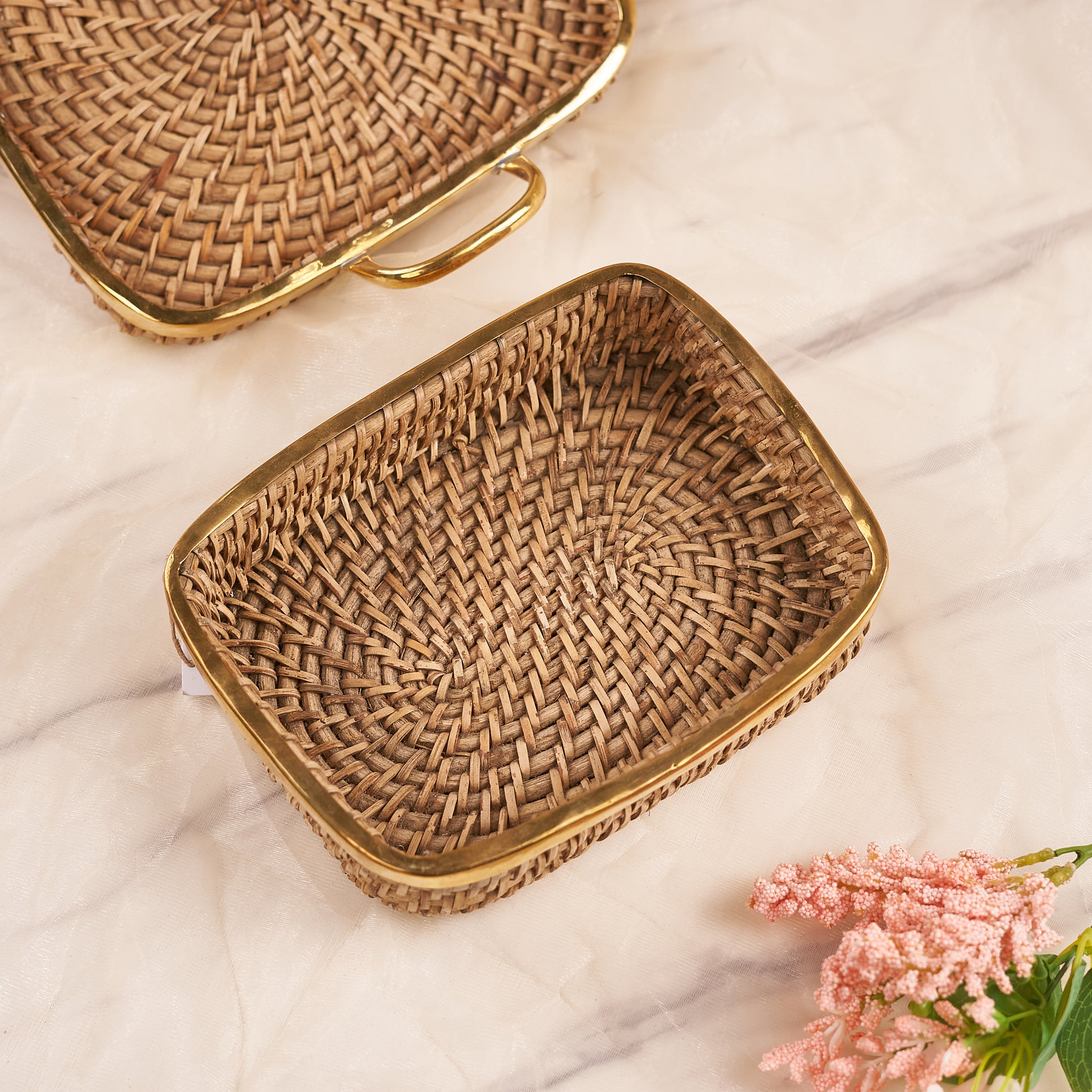 Two Square Rattan Trays with a woven pattern, one with handles visible on the top edge, placed on a marble surface with a bunch of flowers at the bottom right corner.