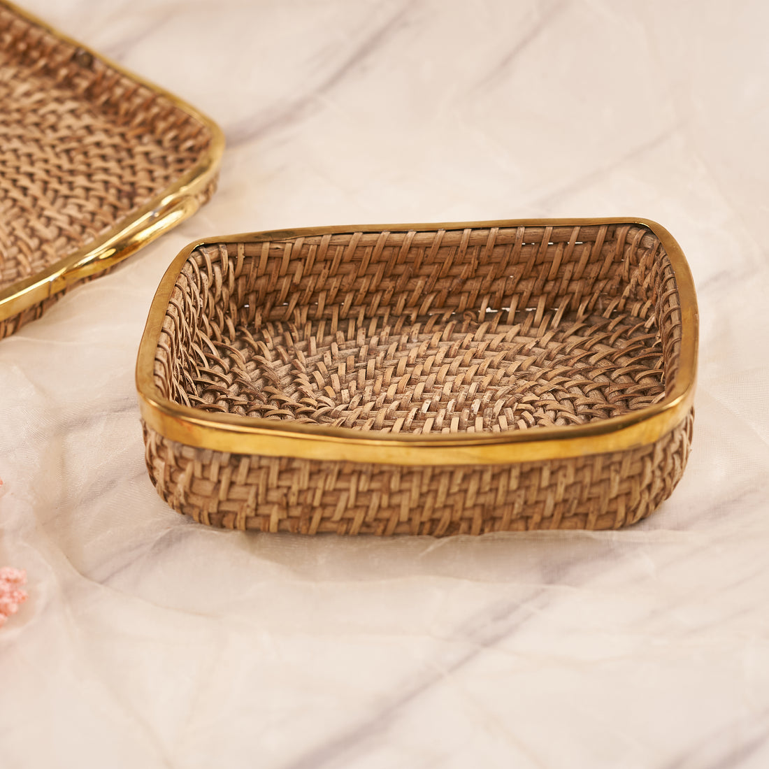 Two Square Rattan Trays with a woven pattern, one with handles visible on the top edge, placed on a marble surface with a bunch of flowers at the bottom right corner.