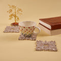 A set of four white wooden carved coasters with an intricate leaf design. A white ceramic cup with colorful heart decals sits on one of the coasters, next to a stack of books and a small decorative tree.