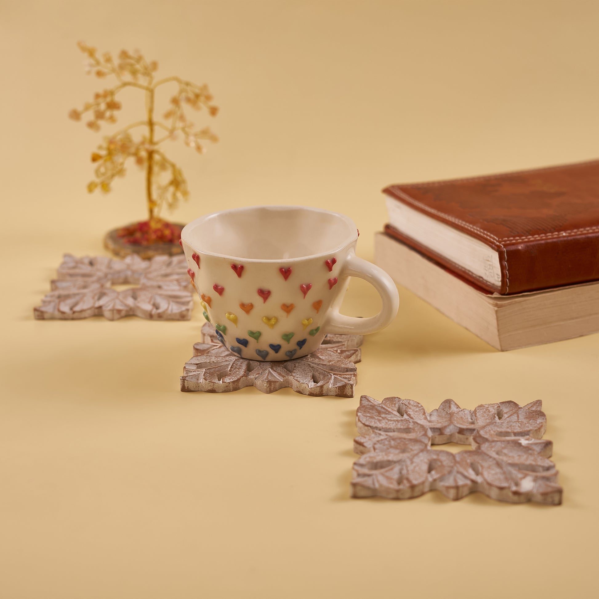 A set of four white wooden carved coasters with an intricate leaf design. A white ceramic cup with colorful heart decals sits on one of the coasters, next to a stack of books and a small decorative tree.