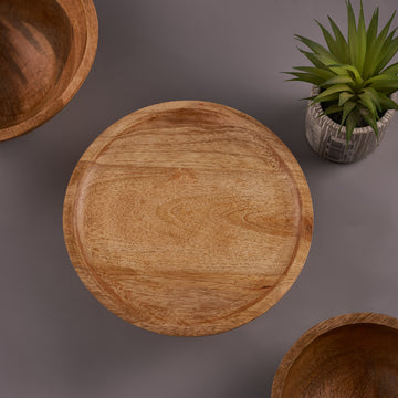 A mango wood cake stand displaying a cake, with a smaller stand visible in the background and a potted plant to the side.