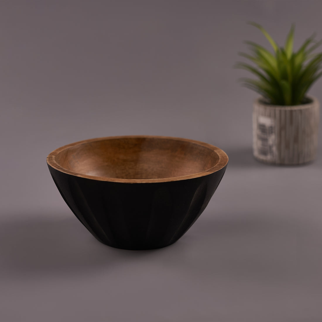 A black Mango Wood bowl with a walnut finish on a gray background, with a plant decoration in the corner.
