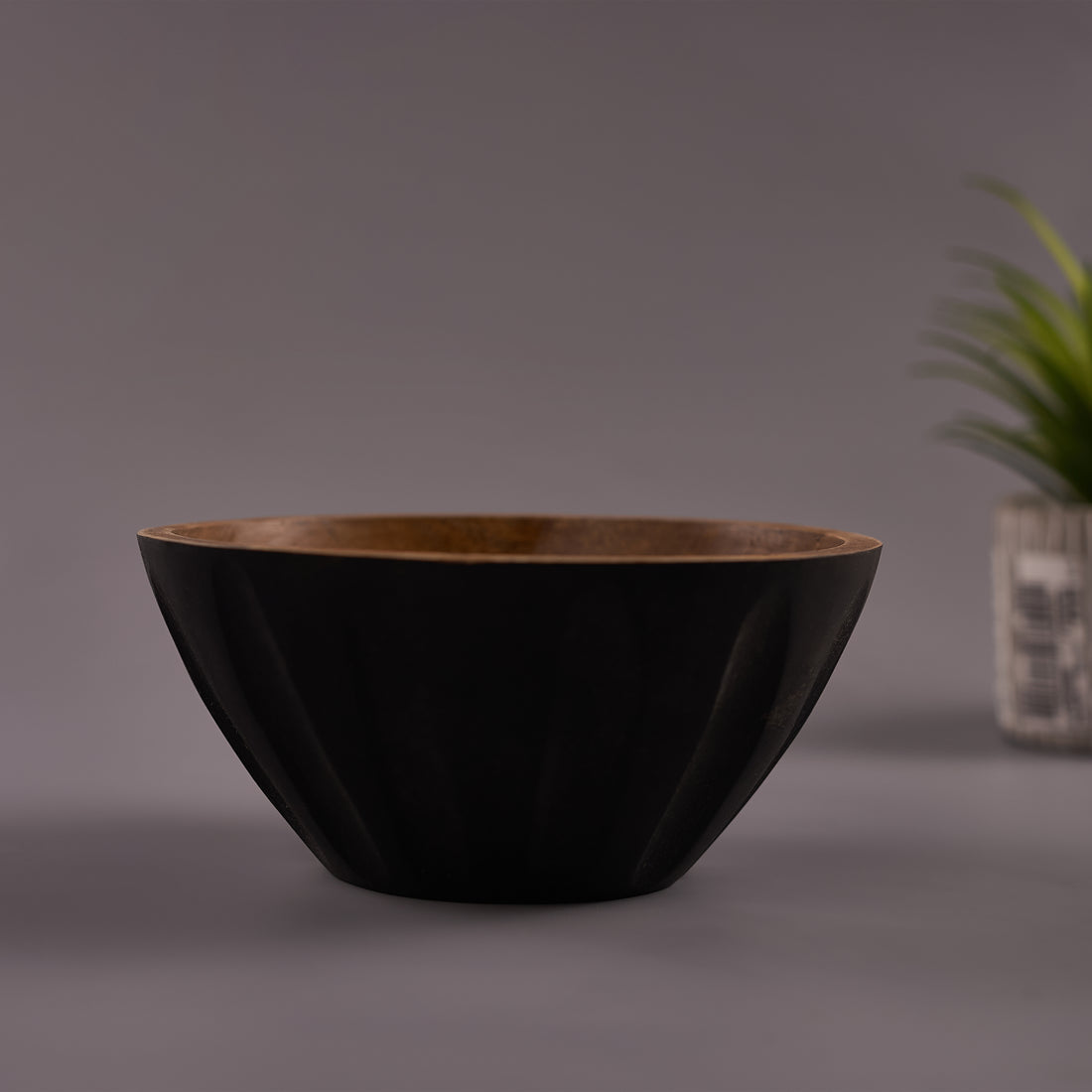 A black Mango Wood bowl with a walnut finish on a gray background, with a plant decoration in the corner.