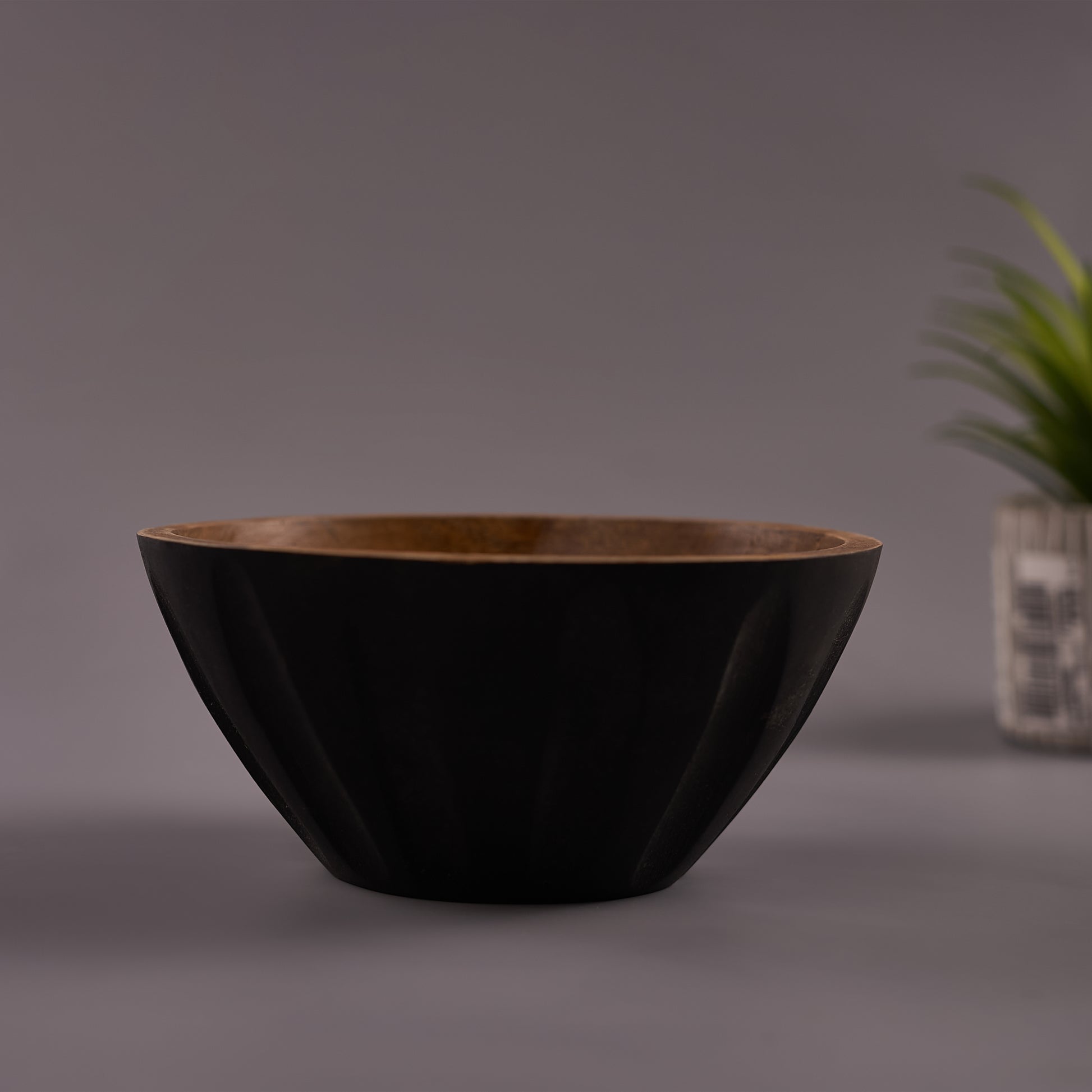 A black Mango Wood bowl with a walnut finish on a gray background, with a plant decoration in the corner.