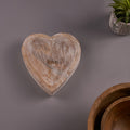 A heart-shaped wooden decor with a rustic finish, placed on a flat surface with a plant in the background.